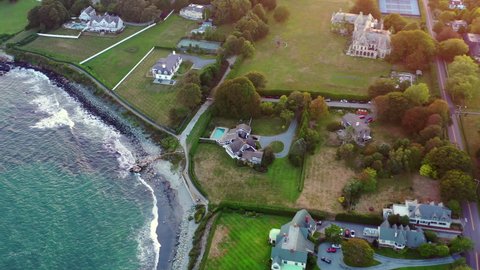Aerial panning shot of houses by waves splashing on coastline, drone flying forward over city at sunset - Newport, Rhode Island