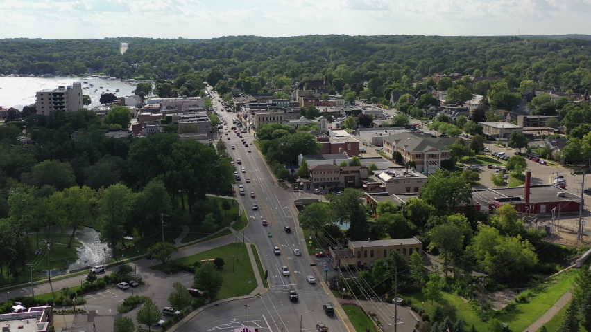 Aerial tilt down shot of vehicles on street amidst buildings in city, drone flying forward over structures by lake against sky - Lake Geneva, Wisconsin