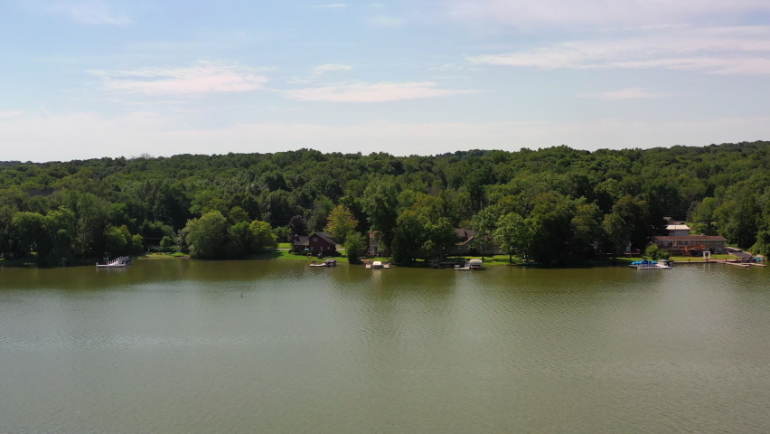 Aerial panning shot of boats and houses amidst green trees at lakeshore, drone flying over lake against sky on sunny day - Lake Como, Wisconsin