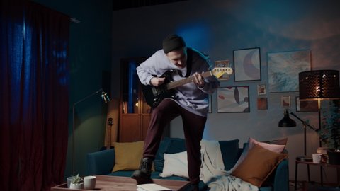 young musician man singing and playing guitar happy and crazy having fun jumping on home sofa couch listening to music and dancing, social distance, band guitarist on lockdown, streaming concert