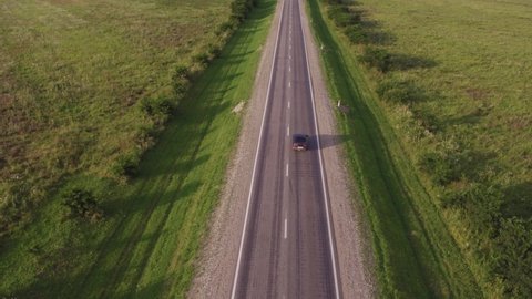 One black sedan car driving on asphalt road through green hilly grass field wih rocky mountains background. Aerial drone wide shot at summer sunny day