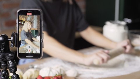 professional culinary blogger performs homemade treats preparation process kneading raw dough with rolling pin while record video. Selective focus on smartphone screen on foreground 