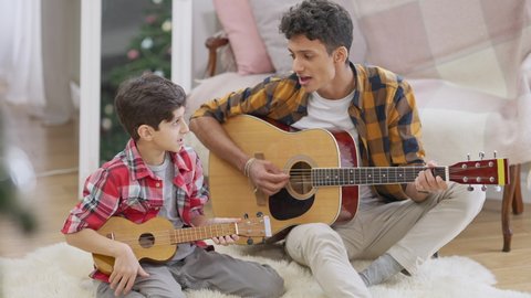Positive Middle Eastern brothers playing guitar and ukulele in slow motion singing song. Wide shot of smiling teenager and child practicing music at home together. Art and lifestyle concept.