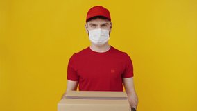 Safety and delivery. Courier in protective mask and gloves wearing red uniform offering parcel box to camera, orange studio background