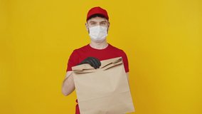 Food order. Young delivery man in red uniform wearing protective medical mask offering eco paper parcel to camera, orange studio background