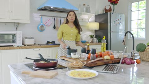 A beautiful Asian girl makes breakfast by looking at how to make it with her laptop computer with a happy face. At her own house