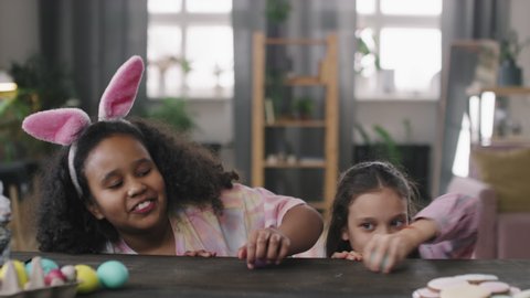 Medium shot of cheerful girls peeping over wooden table and stealing Easter eggs