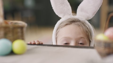 Close up of adorable little boy wearing bunny ears headband peeping at camera from behind table with basket and dyed Easter eggs