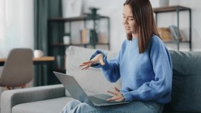 Positive young woman in blue sweater and jeans talking and gesturing during video call on wireless laptop. Happy brunette having online conversation while sitting on couch at home.