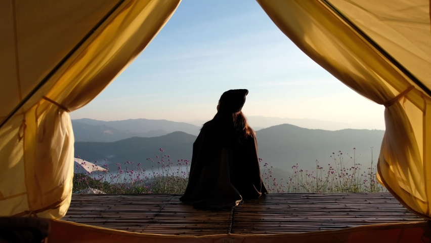 Rear view of a woman sitting on wooden balcony while watching sunrise and a beautiful mountains view outside the tent