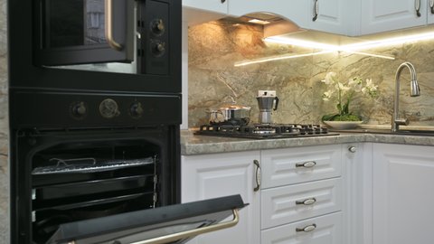 Closeup panning of modern classic luxury white kitchen interior with microwave and electric ovens, gas stove and water tap, ovens doors are open
