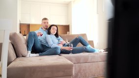 Cheerful young couple, boyfriend and girlfriend watching TV sitting on sofa and drinking wine. Family man and woman spending leisure time together enjoying watching television at home in new apartment