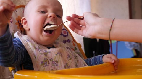 mother's hand feeding baby with a spoon at the high yellow chair while little cute boy looks back and then watching in camera 4K slow mo