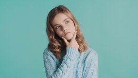 Thoughtful blond teenager girl looking worry keeping hand on chin thinking about something over blue background. Face expression
