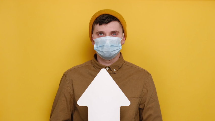 Hesitant man in medical mask hold white arrow points sideways at upper right and left corners, chooses between two options, guy shrug shoulders, looks uncertain, isolated on yellow studio background