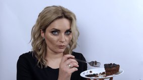 Beautiful young woman model funny eating brownie chocolate cake on white background. Young girl with beautiful smoky eye makeup and black evening dress. Speed video.