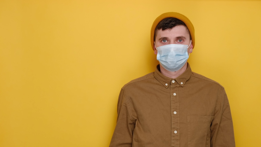 Young caucasian surprised man 20s in medical protective blue mask and gloves hold white arrow, point aside on copy space area mock up isolated on yellow background. People lifestyle, covid 19 concept