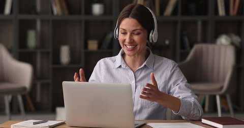 Happy young attractive female entrepreneur in headphones, looking at computer screen, holding distant business negotiations meeting with colleagues or partners using computer video call zoom app.