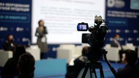 The video camera is filming people sitting on chairs in a conference room. Blurred background with people. Business concept.