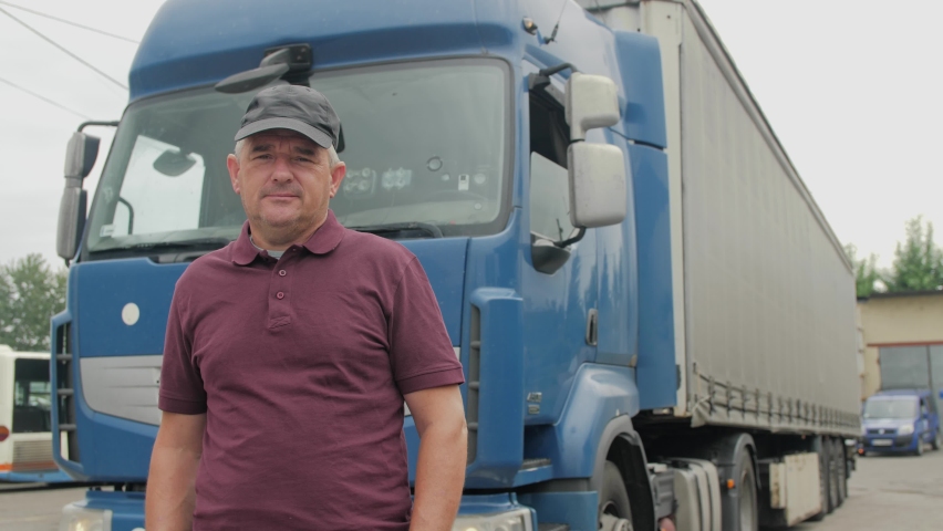 Professional Truck Driver Stands In Front Of Semi Truck with Cargo Trailer Attached. Professional Truck Driver Crossed Arms. In the background is Lorry. 