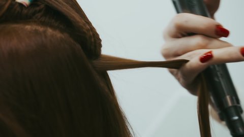Hairdresser with a red manicure on the nails makes a hairstyle for a woman with brown hair. The master selects a strand of hair and makes a corrugation with a curling iron straightener