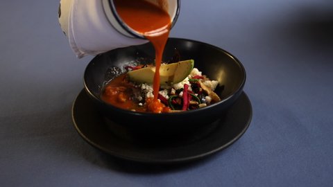 Slowly pouring hot red sauce into black bowl with pork rinds, colorful tortilla slices, and avocado slice. Smooth view of spicy vegetable appetizer above blue tablecloth. Traditional Mexican cuisine
