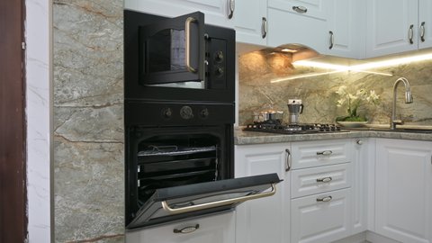 Closeup panning of modern classic luxury white kitchen interior with microwave and electric ovens, gas stove and water tap, ovens doors are open