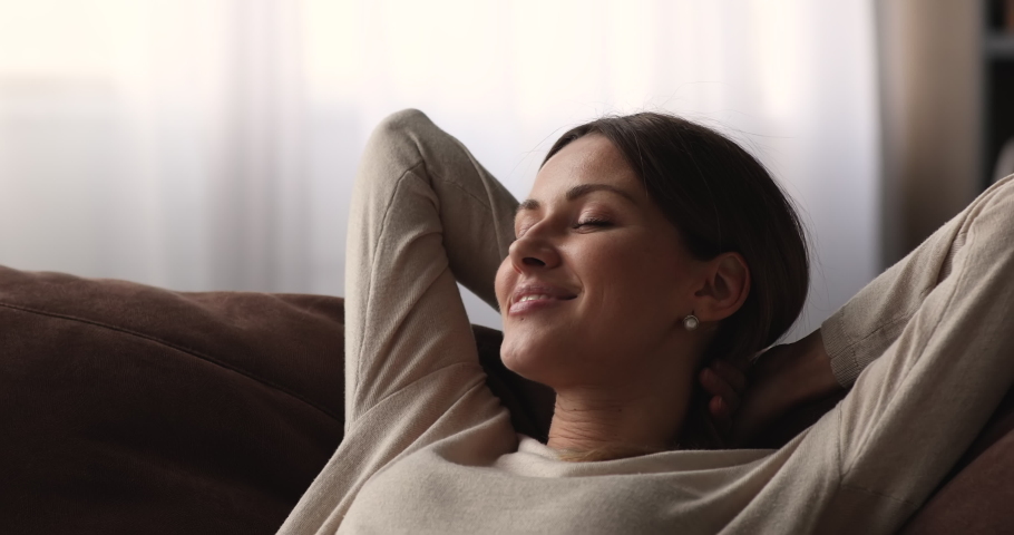 Close up head shot smiling young attractive caucasian woman daydreaming on cozy sofa with folded hands behind head, enjoying peaceful weekend moment alone in living room, lazy weekend pastime. | Shutterstock HD Video #1068565439