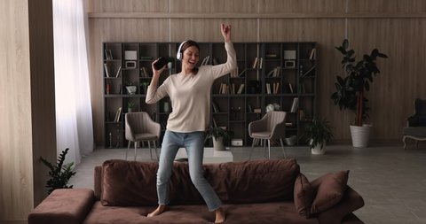 Full length overjoyed young woman in wireless headphones dancing barefoot, jumping on cozy couch in living room, choosing favorite energetic disco music in mobile application, entertaining indoors.
