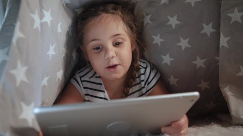 little girl under the blanket with digital tablet. kid dream online video games at network concept. daughter kid watching online video under covers with digital tablet. child on social night media