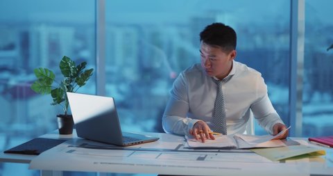 Asian young businessman sitting in corporate cozy office department and doing buisness tasks solving financial issues performing paperwork.