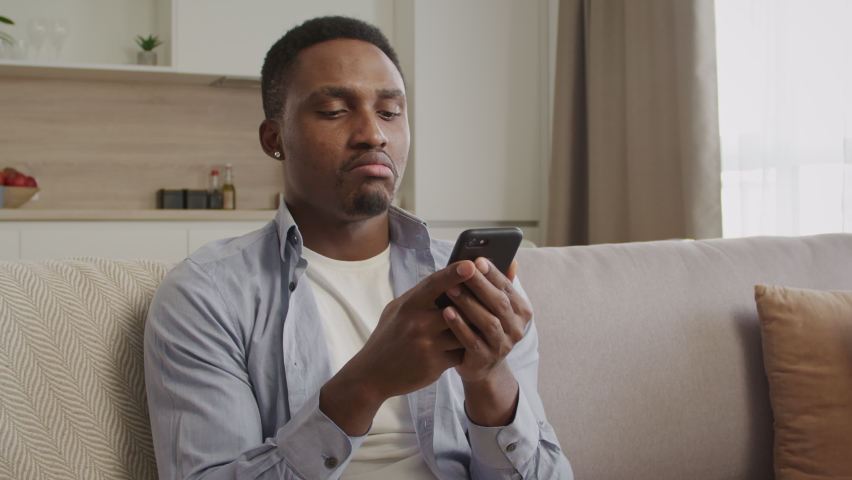 Portrait of a young african american man looking away, thinking and then typing an answer on his smart phone indoors in an apartment | Shutterstock HD Video #1068579056