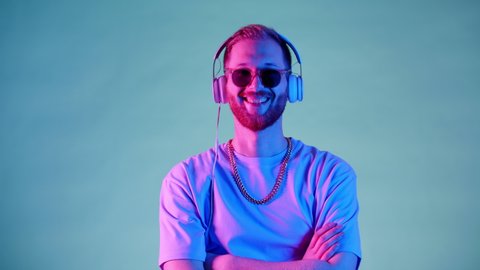 Happy Hipster Young Man With Headphones And Sunglasses Neon Light Studio. Smiling Beautiful Model Standing Alone Looking At Clouse-Up Portrait Camera. Slow motion. Electronic Music Concept