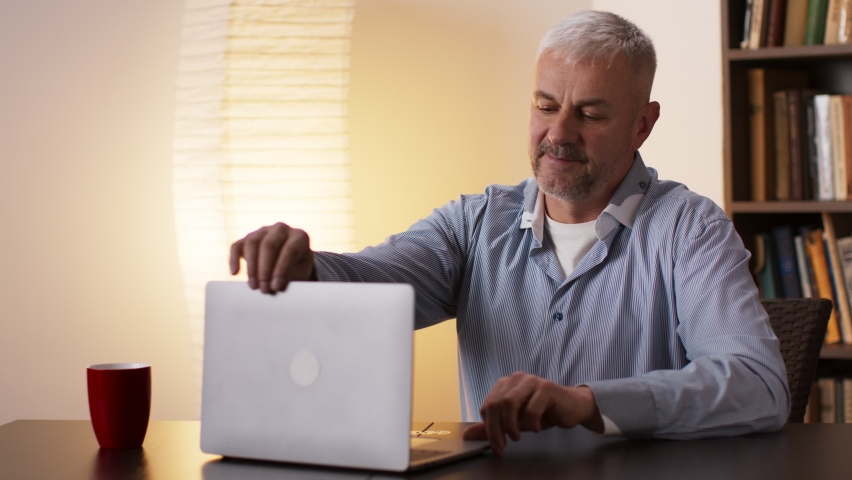 End of working time. Middle aged grey-haired man waving hand to laptop, saying goodbye to colleagues and closing computer, sitting at home office, slow motion Royalty-Free Stock Footage #1068588107