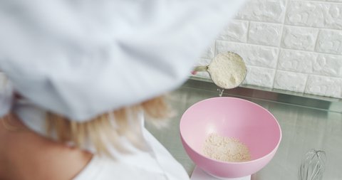 Close up of unrecognizable woman confectioner in white apron, weighing ingredients for cookies, putting almond flour into the pink plastic bowl on electronic weight. mixing dough for macarons