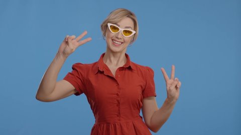 The woman wears her glasses and makes a victory sign (V). Blue studio background. The woman is wearing glasses and a red dress.