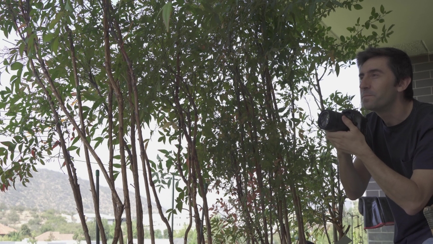 Young man taking pictures of trees with dslr camera. Photographer looking for best angle at home terrace