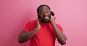 Positive African American man moves with rthythm of music smiles broadly dressed in casual t shirt wears wireless headphones isolated over pink background. People lifesrtyle happiness concept.