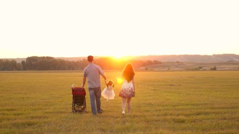 Happy family, child plays with dad and mom on field in light of sun. Little daughter plays and jumps holding hands of dad and mom in park at sunset. Walking with a small child in nature, baby carriage