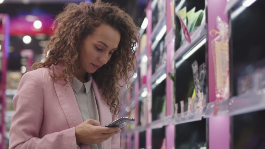 Low-angle medium closeup with slowmo of pretty young woman with long curly hair shopping in cosmetics store scanning qr-code from bottle at smartphone to learn more about product Royalty-Free Stock Footage #1068597386