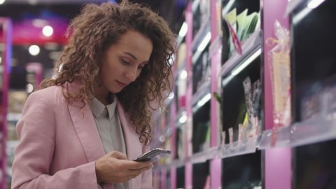 Low-angle medium closeup with slowmo of pretty young woman with long curly hair shopping in cosmetics store scanning qr-code from bottle at smartphone to learn more about product