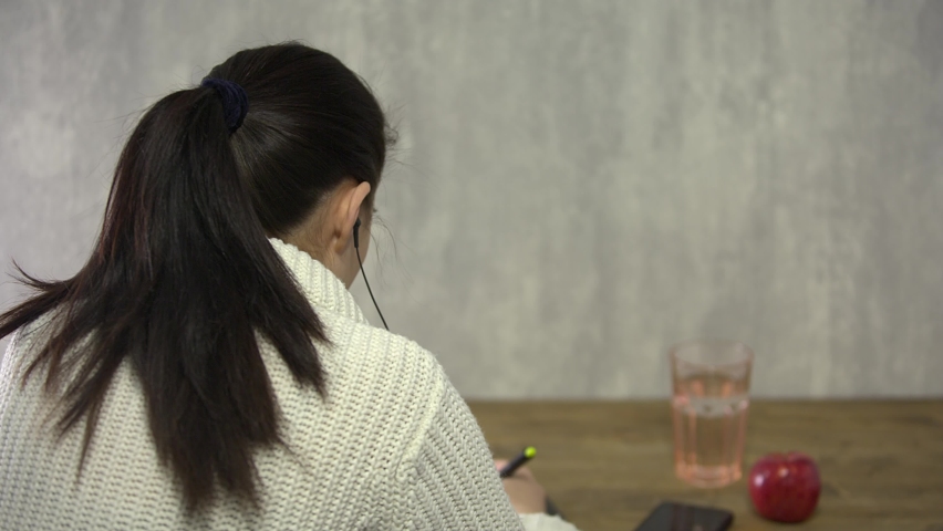 A girl draws on a graphic tablet with a pen, sitting at a wooden table, next to a red apple and a mobile phone. Close-up, selective focus