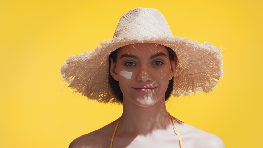 Young good-looking brunette caucasian woman in a straw hat puts spf cream on her cheeks, enjoys the sun and smiles for the camera against yellow background | Spf cream concept | Shutterstock HD Video #1068598766