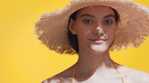 Close-up beauty portrait of young gorgeous dark-haired Сaucasian woman in a straw hat puts sun cream on her cheek, enjoys the sun and smiles at camera against yellow background | Sunscreen commercial