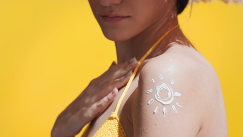 Young slim dark-haired woman in a yellow crochet swimwear and a straw hat puts sunscreen on her shoulder against yellow background | Body care concept