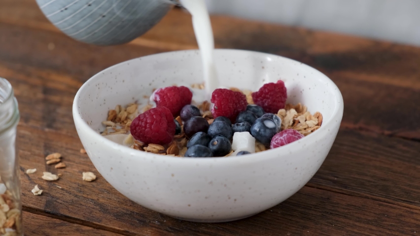Milk pouring in granola bowl with fresh blueberries and raspberries on a wooden table. Healthy breakfast food, cereals with milk | Shutterstock HD Video #1068603266