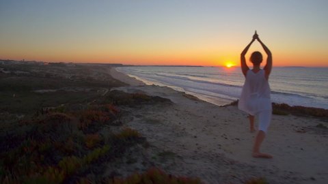 DRONE, LENS FLARE: Cinematic shot of a flexible young woman performing yoga on top of a cliff facing the endless ocean. Female tourist does relaxing yoga on the beach of Peniche at golden sunset.