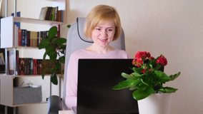 Caucasian young woman sits at laptop and types in cozy room. Nearby on table is houseplant. Behind - bookcase. Concept of remote work from home, lockdown, freelance, online training, stay home
