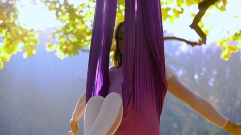 Beautiful athletic young woman doing air yoga on hammock in nature, complex acrobatic exercises in a park.
Fit girl changes poses in air, practicing and breathing in the sunlight.