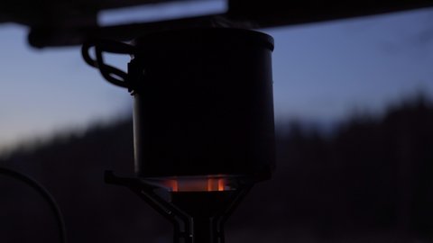 Tourist pot being heated at camping gas stove at twilight. Preparing hot drink during outdoor adventure.